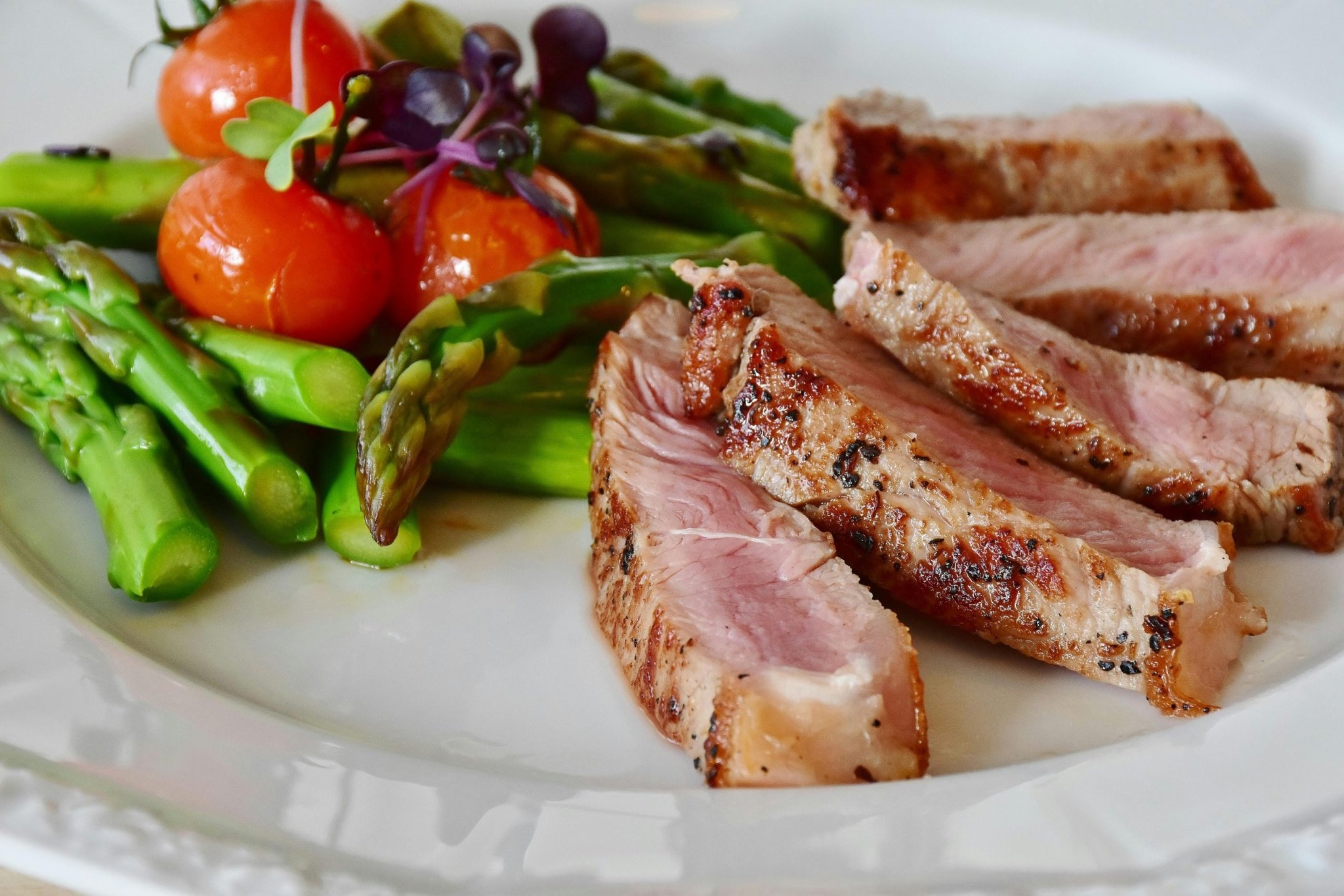 Grilled steak slices served with asparagus, cherry tomatoes, and microgreens on a white plate